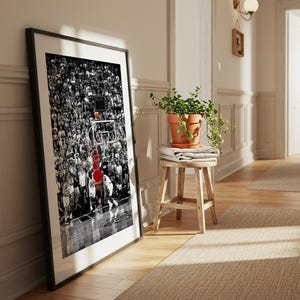 May include: A framed vertical wall art print featuring a black and white photograph of a crowded basketball arena. A single player in a bright red jersey is captured in motion near the hoop, providing a stark color contrast to the monochrome scene.