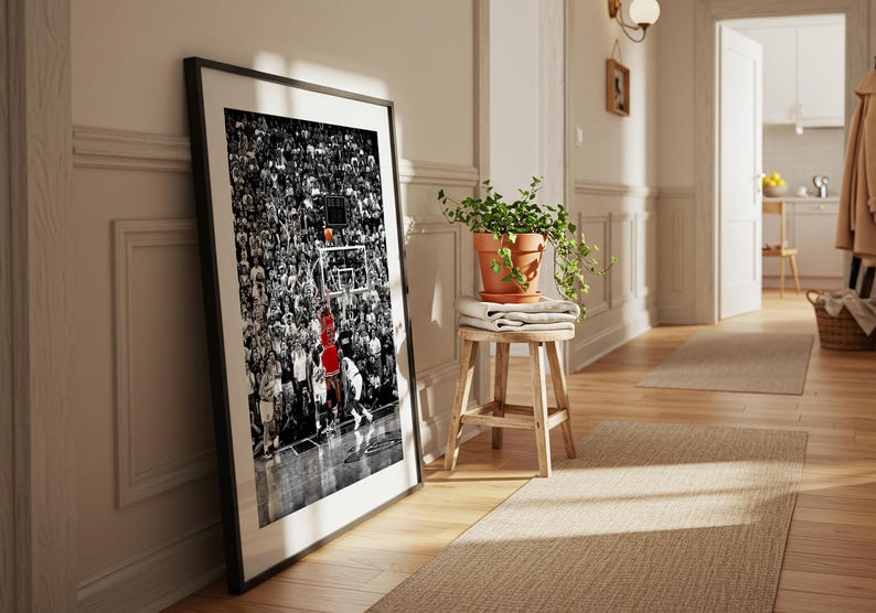 May include: A framed vertical wall art print featuring a black and white photograph of a crowded basketball arena. A single player in a bright red jersey is captured in motion near the hoop, providing a stark color contrast to the monochrome scene.