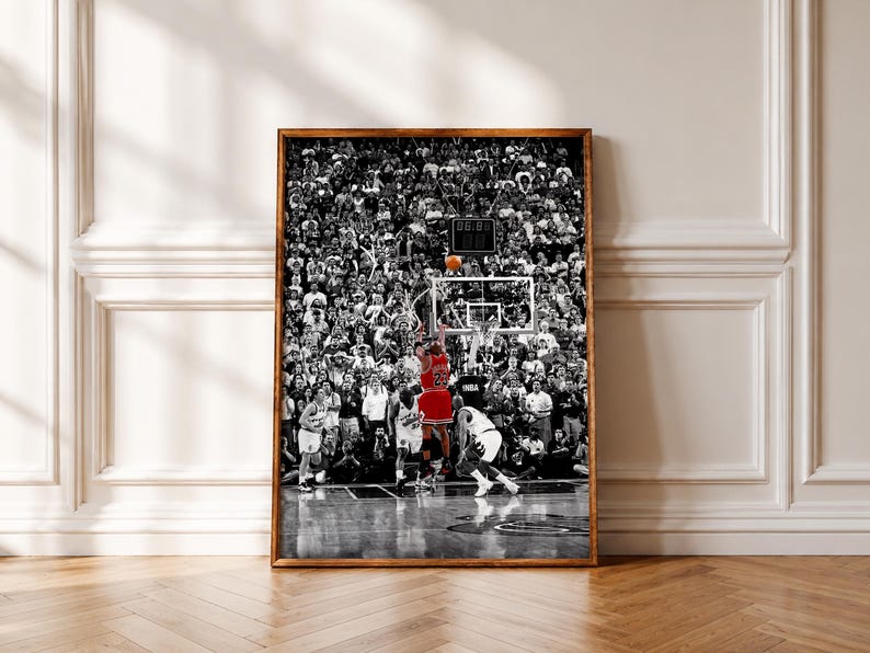 May include: Black and white framed print of a basketball game. A player in a red jersey with the number 23 is shooting a basketball. The crowd is in the background, and the scoreboard shows the time. The image is in a black frame.