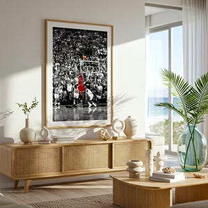 May include: A framed vertical wall art print featuring a black and white photograph of a basketball game with a player in a red jersey shooting a basketball toward the hoop, displayed above a light wood media console with cane cabinet doors.
