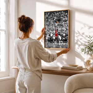 May include: A framed rectangular wall art print featuring a black and white photograph of a crowded basketball arena. A single player in a bright red jersey is captured in the center, jumping to shoot a basketball toward the hoop.