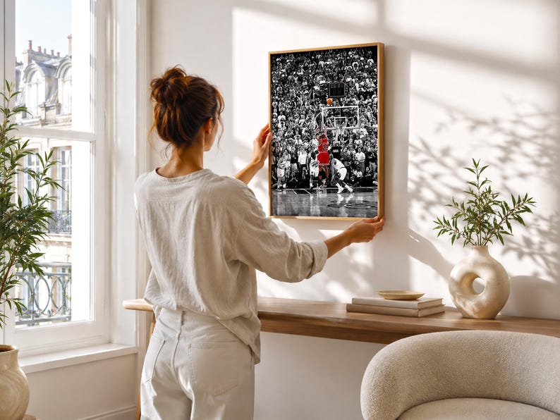 May include: A framed rectangular wall art print featuring a black and white photograph of a crowded basketball arena. A single player in a bright red jersey is captured in the center, jumping to shoot a basketball toward the hoop.