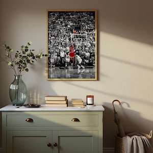 May include: A framed rectangular wall art print featuring a black and white photograph of a basketball game with a player in a red jersey jumping toward the hoop. The print is displayed in a thin light wood frame hanging on a neutral wall.