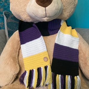 May include: A crocheted scarf with purple, white, yellow and black stripes. The scarf has tassels on the ends and a button on one side.