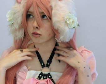 May include: A pink wig with long straight hair, featuring fluffy white animal-style ear attachments adorned with small pink strawberry accents, white fabric roses, and a glittery pink bow on top. A black leather-like harness with a metal ring.