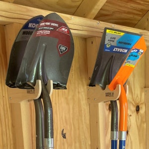 May include: Three shovels are mounted on a wooden wall. Two shovels have dark gray blades and black handles. The third shovel has an orange handle and a dark gray blade. The shovels are labeled with the brand name and product information.