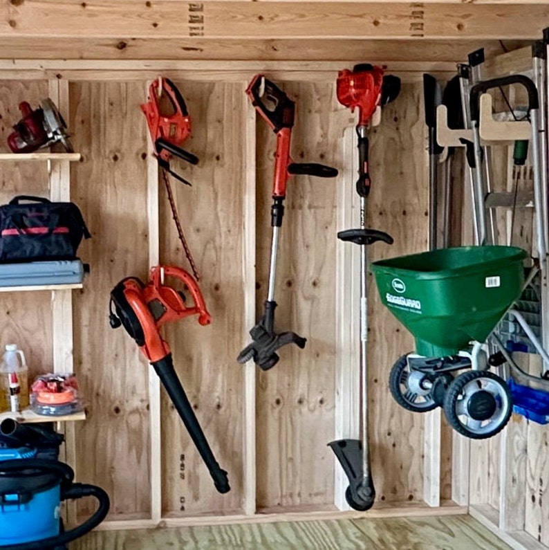 May include: A wall-mounted storage system for gardening tools.  The system includes hooks for hanging tools, a shelf for storing smaller items, and a green plastic spreader with a white handle.  The tools include a red and black leaf blower, a black and red string trimmer, a black and red weed trimmer, and a red and black hedge trimmer.