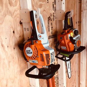 May include: Two orange and white Stihl hedge trimmers are hanging on a wooden wall. The trimmers are attached to wooden wall mounts with the words "UP" printed on them. The trimmers have black handles and black blades.
