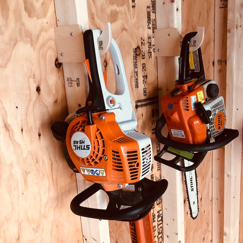 May include: Two orange and white Stihl hedge trimmers are hanging on a wooden wall. The trimmers are attached to wooden wall mounts with the words "UP" printed on them. The trimmers have black handles and black blades.
