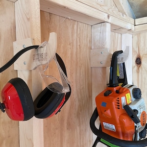 May include: A wooden wall with two hooks mounted on it. The hooks are labeled "Hang This Up" and are used to store a pair of red and black earmuffs and a chainsaw.