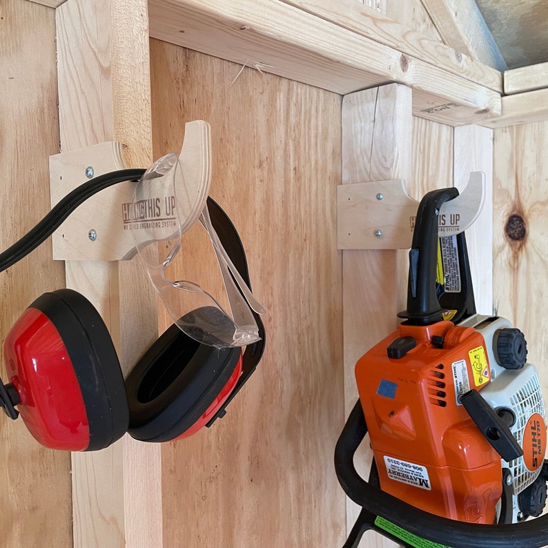 May include: A wooden wall with two hooks mounted on it. The hooks are labeled "Hang This Up" and are used to store a pair of red and black earmuffs and a chainsaw.