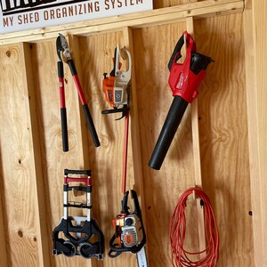 May include: A wooden wall with a variety of tools hanging on it. The tools include a leaf blower, a hedge trimmer, a chainsaw, and a hand rake. The wall is painted a light brown color and has a sign that says "My Shed Organizing System".
