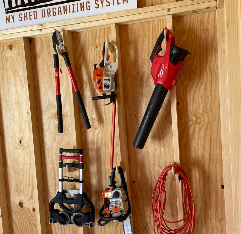 May include: A wooden wall with a variety of tools hanging on it. The tools include a leaf blower, a hedge trimmer, a chainsaw, and a hand rake. The wall is painted a light brown color and has a sign that says "My Shed Organizing System".