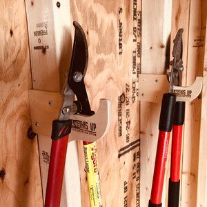 May include: Two red-handled gardening tools are hanging on wooden wall mounts. The tools are labeled "ANGTHIS UP" and are attached to the wall with screws. The wall is made of plywood and has some writing on it.