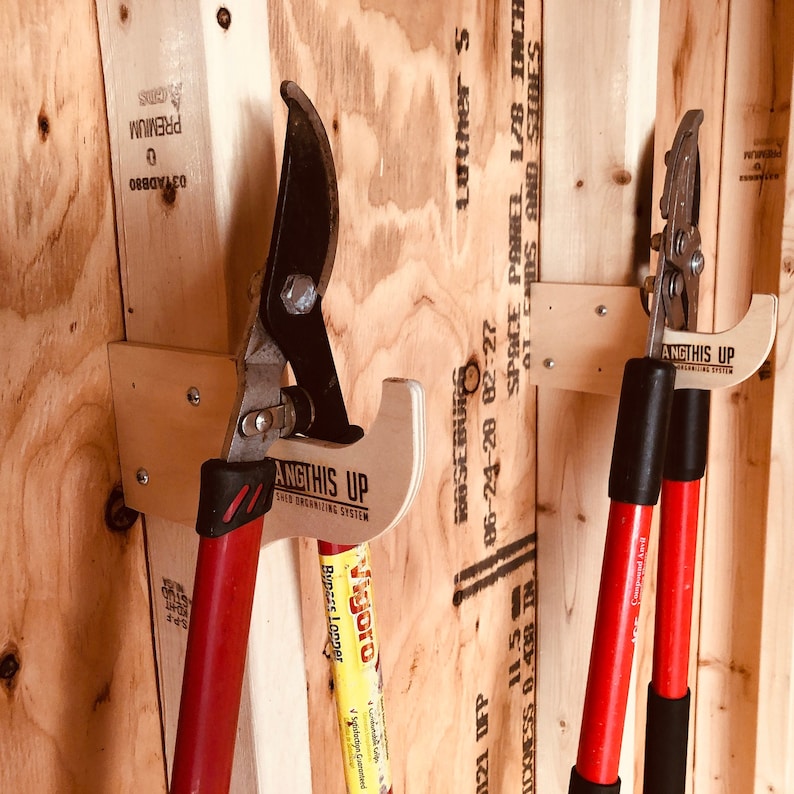 May include: Two red-handled gardening tools are hanging on wooden wall mounts. The tools are labeled "ANGTHIS UP" and are attached to the wall with screws. The wall is made of plywood and has some writing on it.