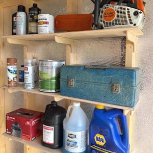 May include: A wooden shelving unit holding various items including a white and orange Stihl MS 192 T chainsaw, spray paint cans, a blue metal toolbox, a red box of trash bags, a black jug of bar and chain oil, and jugs of windshield washer fluid.