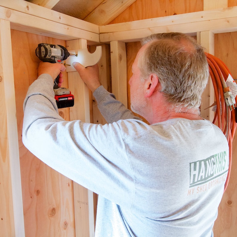 May include: A person uses a power drill to install a white plastic shelf bracket on a wooden wall. The wall is made of wood planks. The person is wearing a grey long-sleeved shirt with the text "Hang This My Shed Outfitters" printed on the back.