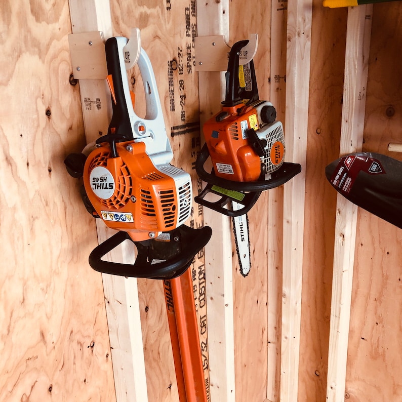 K&ouml;nnte beinhalten: Zwei orangefarbene und schwarze Kettens&auml;gen, die an Holz-Wandhalterungen h&auml;ngen. Die Kettens&auml;gen sind mit "STIHL" beschriftet und haben schwarze Griffe. Die Kettens&auml;gen h&auml;ngen an einer Wand mit Holzverkleidung.