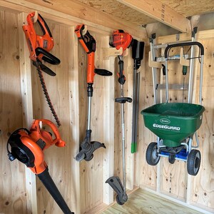 May include: A shed with a variety of gardening tools hanging on the wall. There is a green and black spreader with the text "Scotts EdgeGuard Total Lawn Food" on the side. There are also several orange and black weed trimmers and a black and orange hedge trimmer.