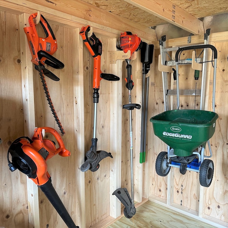 May include: A shed with a variety of gardening tools hanging on the wall. There is a green and black spreader with the text "Scotts EdgeGuard Total Lawn Food" on the side. There are also several orange and black weed trimmers and a black and orange hedge trimmer.