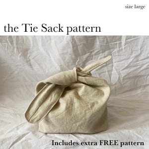 Op de afbeelding: Een beige stoffen zak met een knoop, maat large, met een geknoopt handvat. De tekst "the Tie Sack pattern" staat boven de zak en "Includes extra FREE pattern" staat eronder. De zak is gemaakt van een getextureerd, natuurlijk gekleurd materiaal.