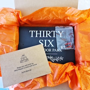 May include: A black slate sign with white text that reads "THIRTY SIX DOOR PARK". The sign is wrapped in orange tissue paper and a small brown card with a gift box icon and the text "Hope you will both be very happy in your new home Love Sam xx" is visible.