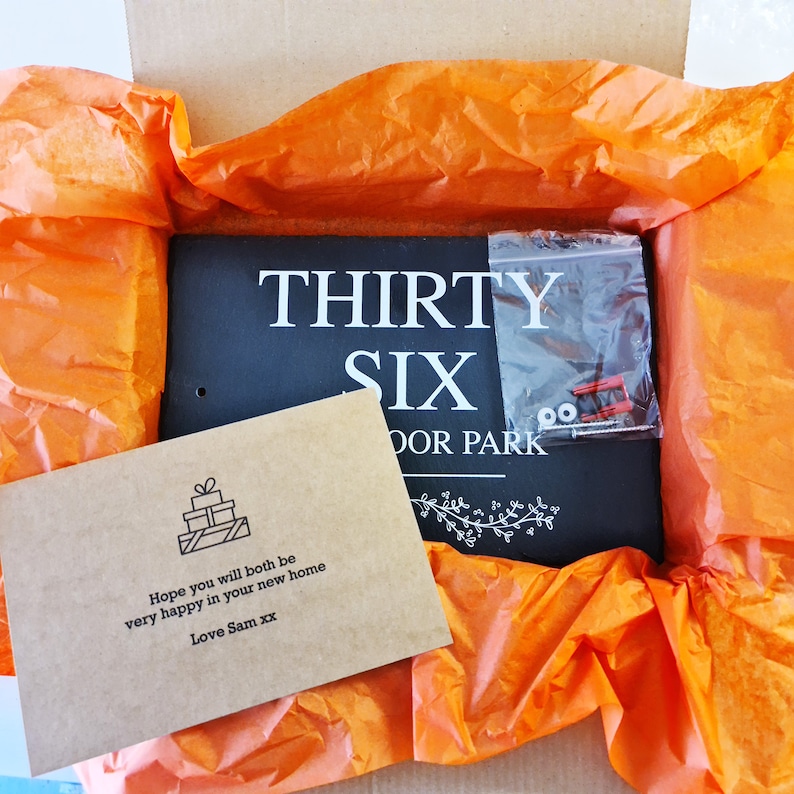 May include: A black slate sign with white text that reads "THIRTY SIX DOOR PARK". The sign is wrapped in orange tissue paper and a small brown card with a gift box icon and the text "Hope you will both be very happy in your new home Love Sam xx" is visible.