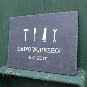Könnte beinhalten: Schwarzes Schiefer-Schild mit weißem Text und Symbolen. Der Text lautet "Dads Werkstatt Est 2017". Die Symbole sind ein Hammer, ein Schraubendreher, eine Säge und ein Schraubenschlüssel.