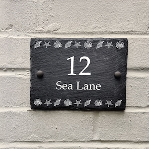 以下が含まれることがあります： 白い文字と装飾的な貝殻が付いた黒いスレート製の家の番号札。番号の12と「Sea Lane」の文字が札に書かれている。