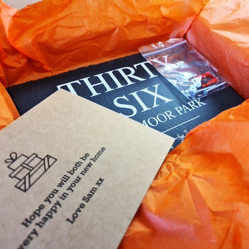 May include: A brown card with the text "Hope you will both be very happy in your new home Love Sam xx" and a small gift box icon. The card is in a box with orange tissue paper and a black slate sign with the text "THIRTY SIX MOOR PARK".