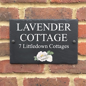 May include: Black slate house sign with white text that reads "Lavender Cottage" and "7 Littledown Cottages". A white flower with a green stem is in the bottom right corner.