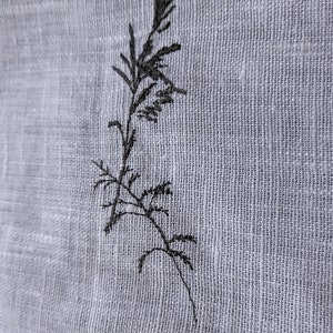 May include: A black embroidered design of a single plant stem with leaves on a white fabric background.