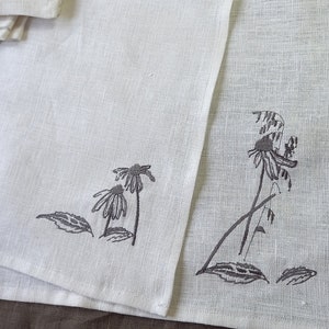 May include: Two white linen tea towels with black embroidered floral designs. One towel features a single flower with a long stem and leaves, while the other towel features three flowers with shorter stems and leaves.
