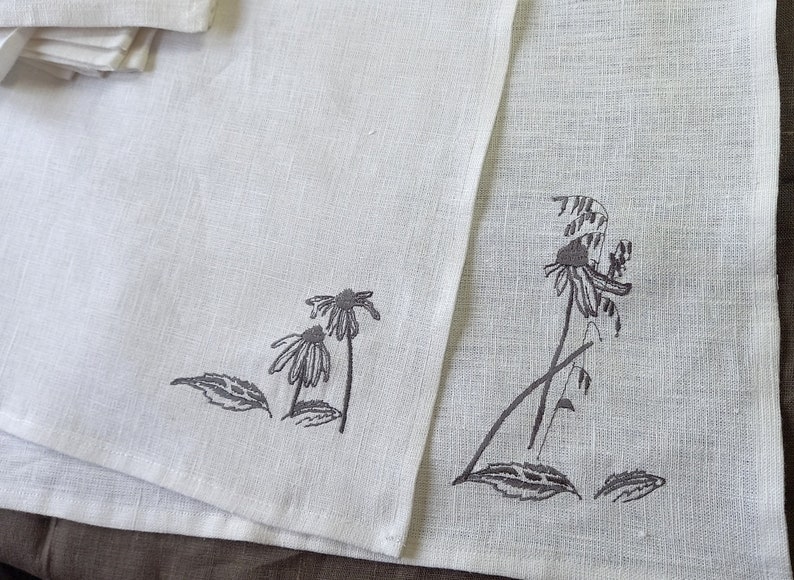 May include: Two white linen tea towels with black embroidered floral designs. One towel features a single flower with a long stem and leaves, while the other towel features three flowers with shorter stems and leaves.