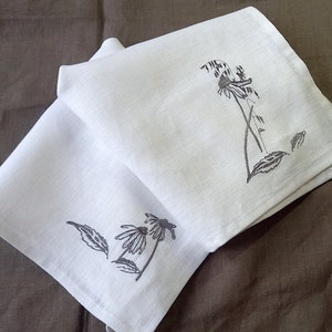 May include: Two white linen tea towels with black embroidered floral designs. The design on the top towel features a single flower with leaves. The design on the bottom towel features two flowers with leaves.