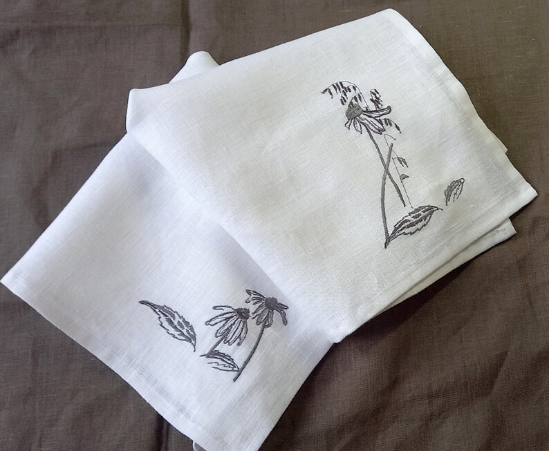 May include: Two white linen tea towels with black embroidered floral designs. The design on the top towel features a single flower with leaves. The design on the bottom towel features two flowers with leaves.