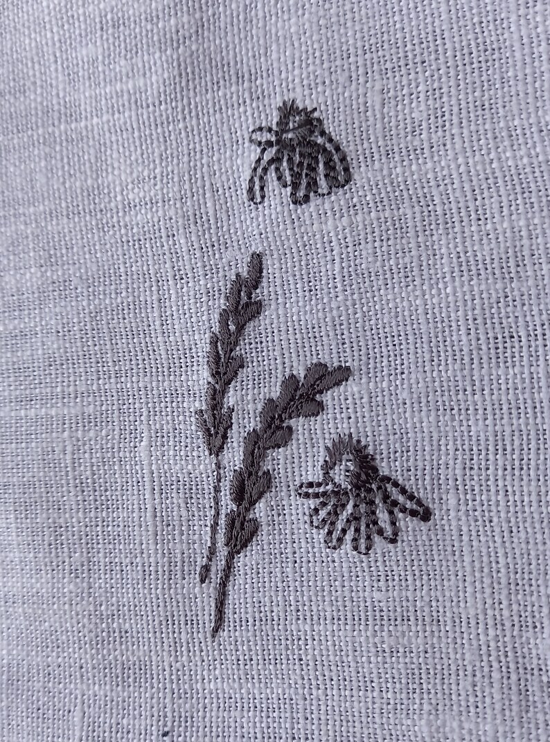 May include: Embroidered design on a white fabric featuring two stylized bees and a sprig of leaves.