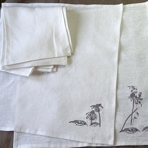 May include: A set of white linen napkins with a gray embroidered design of flowers and leaves. The napkins are folded and stacked on a brown surface.