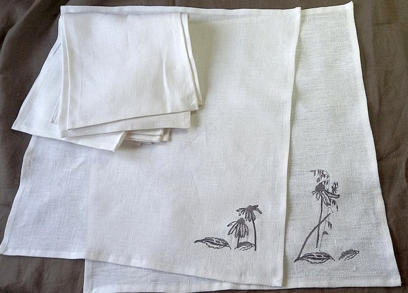 May include: A set of white linen napkins with a gray embroidered design of flowers and leaves. The napkins are folded and stacked on a brown surface.