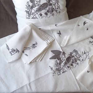 May include: A set of white linen napkins with a black and white bird and floral design. The napkins are folded and stacked on a white tablecloth.