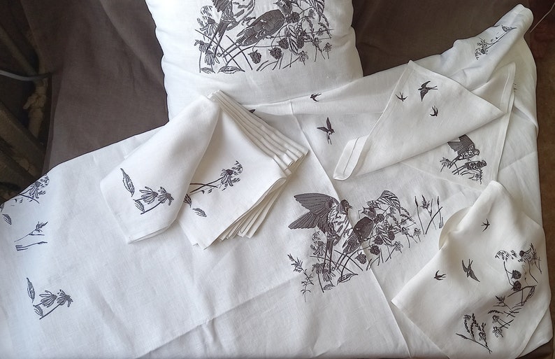May include: A set of white linen napkins with a black and white bird and floral design. The napkins are folded and stacked on a white tablecloth.