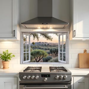 Puede incluir: Una cocina moderna con armarios blancos, una campana extractora de acero inoxidable y una estufa de gas. Una ventana con una vista panorámica de un paisaje se encuentra sobre la estufa. Una pequeña planta en maceta añade un toque de vegetación.