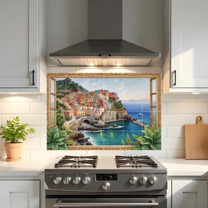 Puede incluir: Un mural de azulejos para la cocina que representa una escena de un pueblo costero italiano. La obra de arte, enmarcada por un borde dorado, muestra edificios coloridos, un puente y el mar. La escena se ve a través de una ventana, con follaje verde en la parte inferior.