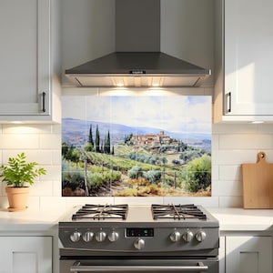 Op de afbeelding: Een keukenachterwand met een landschapsschilderij van een Toscaans dorp. Het kunstwerk toont glooiende heuvels, wijngaarden, cipressen en een verre stad onder een blauwe lucht. De achterwand is geïnstalleerd boven een roestvrijstalen gasfornuis.