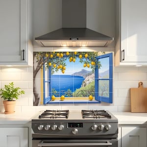 May include: A kitchen backsplash tile mural showing a coastal scene. The mural features a blue window frame with a view of the sea, a lemon tree, and a coastal town. The scene is framed by white cabinets and a stainless steel cooker hood.