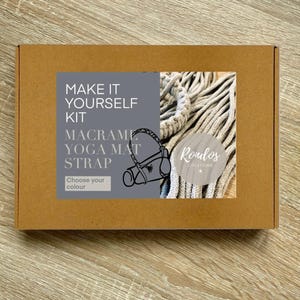 May include: A brown cardboard box with a grey label that says "Make It Yourself Kit Macrame Yoga Mat Strap Choose your colour" and a white macrame yoga mat strap. The label also has a white circle with the text "Rondos Creations" in black.