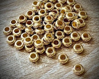 May include: A collection of small, round, metallic gold-toned beads with a smooth, polished finish. Each bead features a central hole, creating a uniform ring-like shape. The beads are scattered in a loose pile, showing their reflective surface.