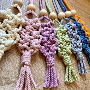 May include: A set of seven macrame keychains with wooden beads and tassels in various colours, including cream, pink, purple, green, blue, and yellow.