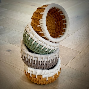 May include: A set of four woven baskets in different colors and patterns. The baskets are stacked on top of each other, with the largest basket at the bottom and the smallest basket at the top. The baskets are made of natural fibers and have a rustic, handmade look.