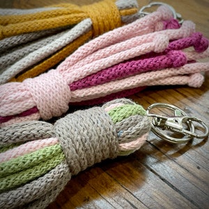 May include: Three colourful keychains made of woven yarn. The keychains are in shades of pink, grey, green, and yellow. Each keychain has a silver metal key ring.